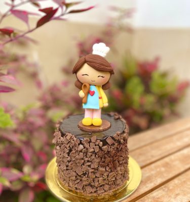 Encantadora mini torta de chocolate decorada con una figura de mamá cocinera y un topper dorado para celebrar el Día de la Madre.