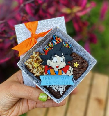 Mini Four Goku decorado con fondant y toppings variados, empaquetado en una caja ecológica con opción de sticker personalizado.