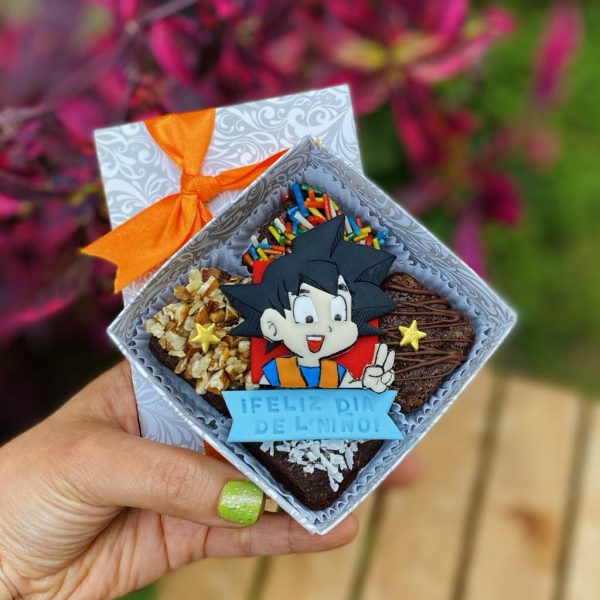 Mini Four Goku decorado con fondant y toppings variados, empaquetado en una caja ecológica con opción de sticker personalizado.